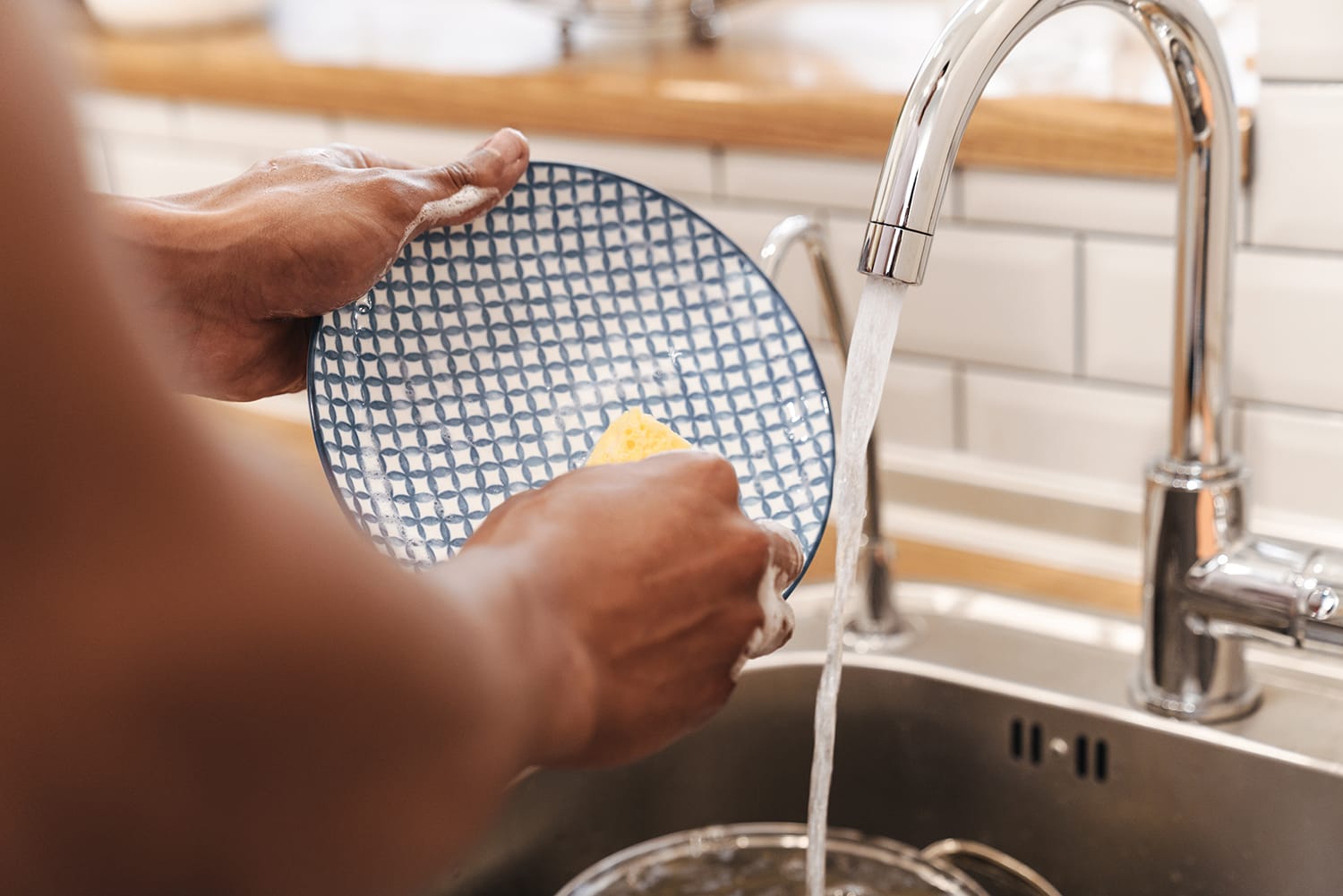 Close-up of a person's hands rinsing a patterned blue and white plate under running water in a kitchen sink.