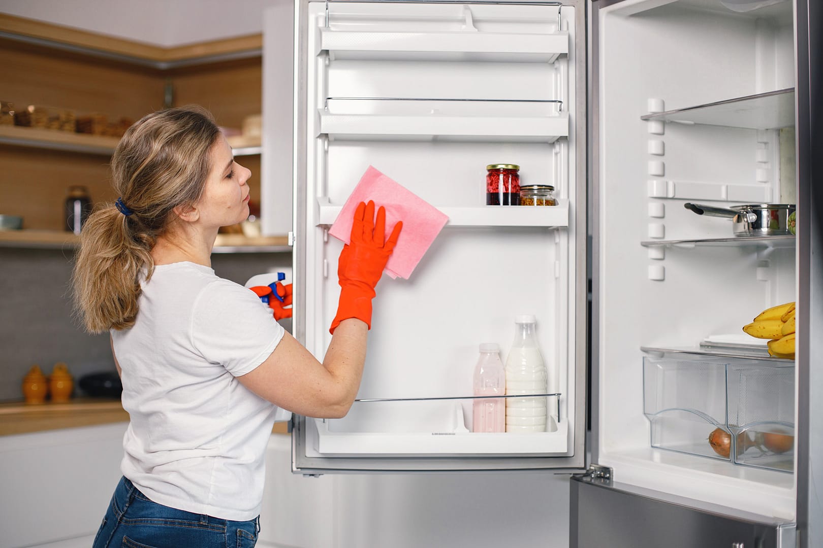 - Sam's Appliance Repair Woman wearing red rubber gloves cleaning the inside of a refrigerator with a pink cloth and spray cleaner in a modern kitchen.
