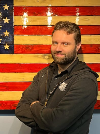 appliance-repairman-nathan Appliance repairman standing in front of an American Flag with his arms crossed looking confident.